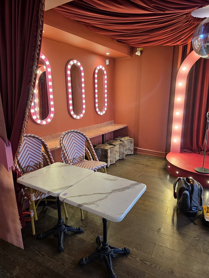 Pink-walled bar room with bulb-framed mirrors, marble tables and a small lit stage