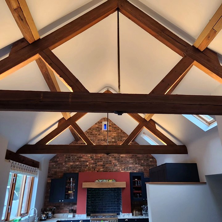 Vaulted ceiling with exposed timber trusses lit by wall uplighters, above a kitchen with a brick chimney breast