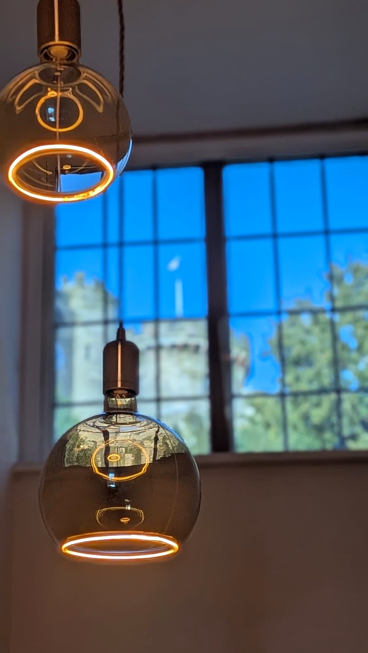 Two large globe filament pendant lights hanging in front of a leaded window, with the walls of Warwick Castle beyond