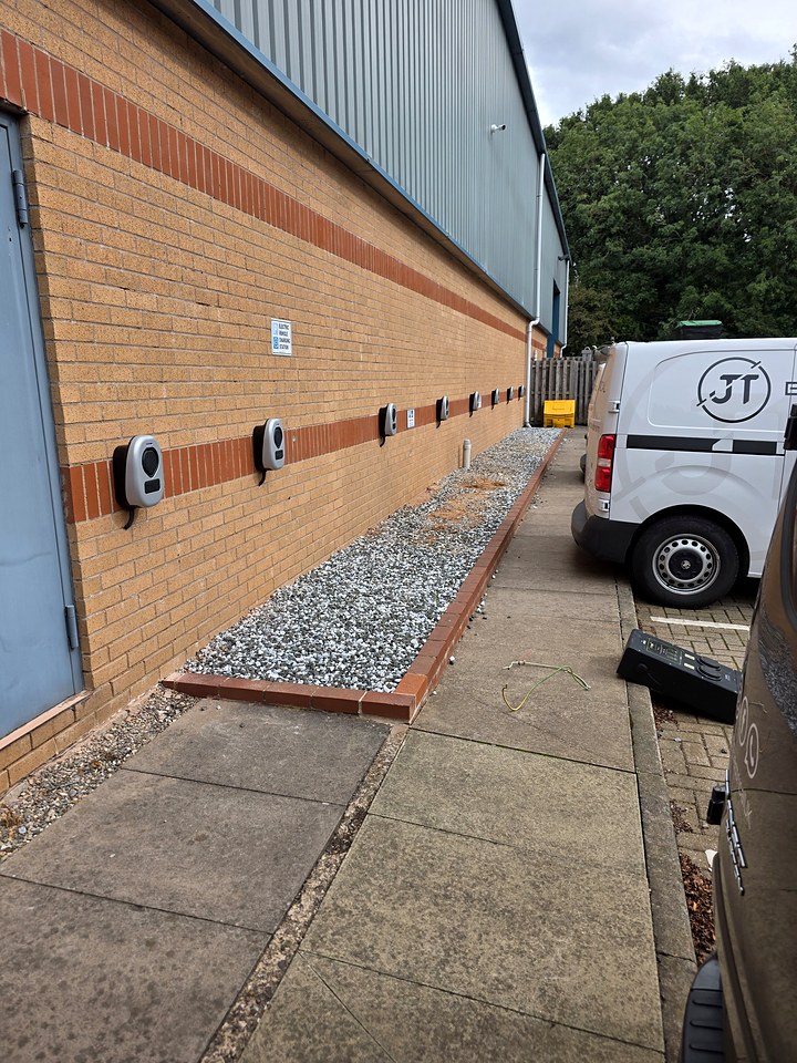 Row of wall-mounted EV chargers along the brick wall of an industrial unit, with a JT Electrical van parked alongside