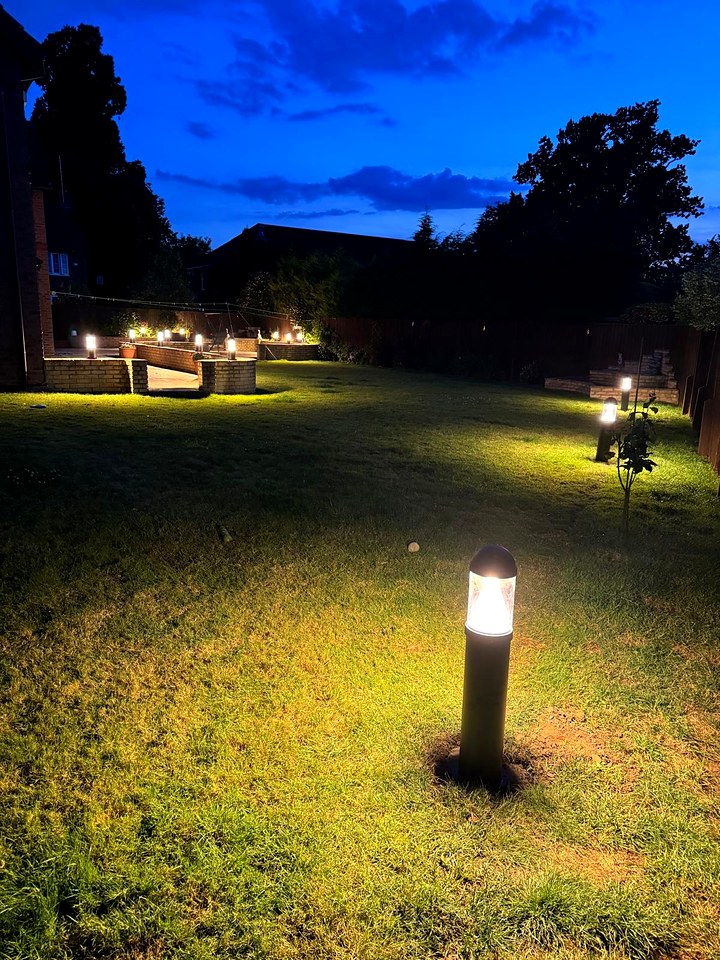Black bollard light glowing on a lawn at dusk, with more garden lights along a low wall behind