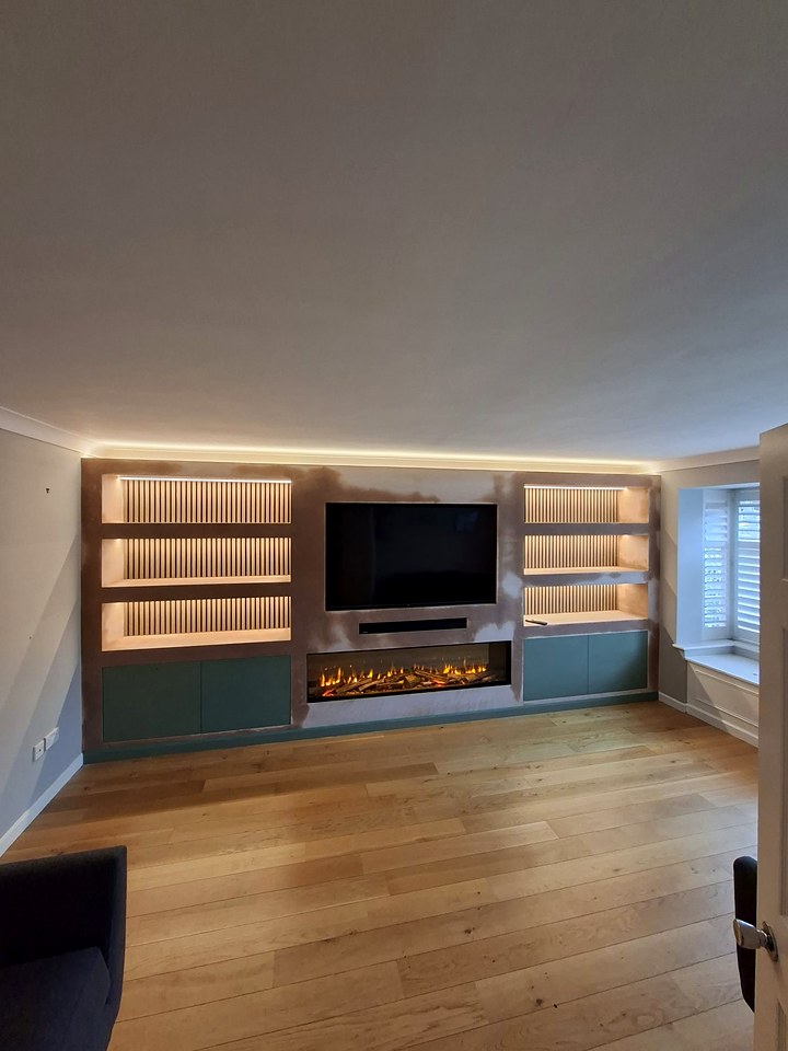 Finished media wall with LED-lit shelves either side of a wall-mounted TV and an electric fire, in a living room with oak flooring