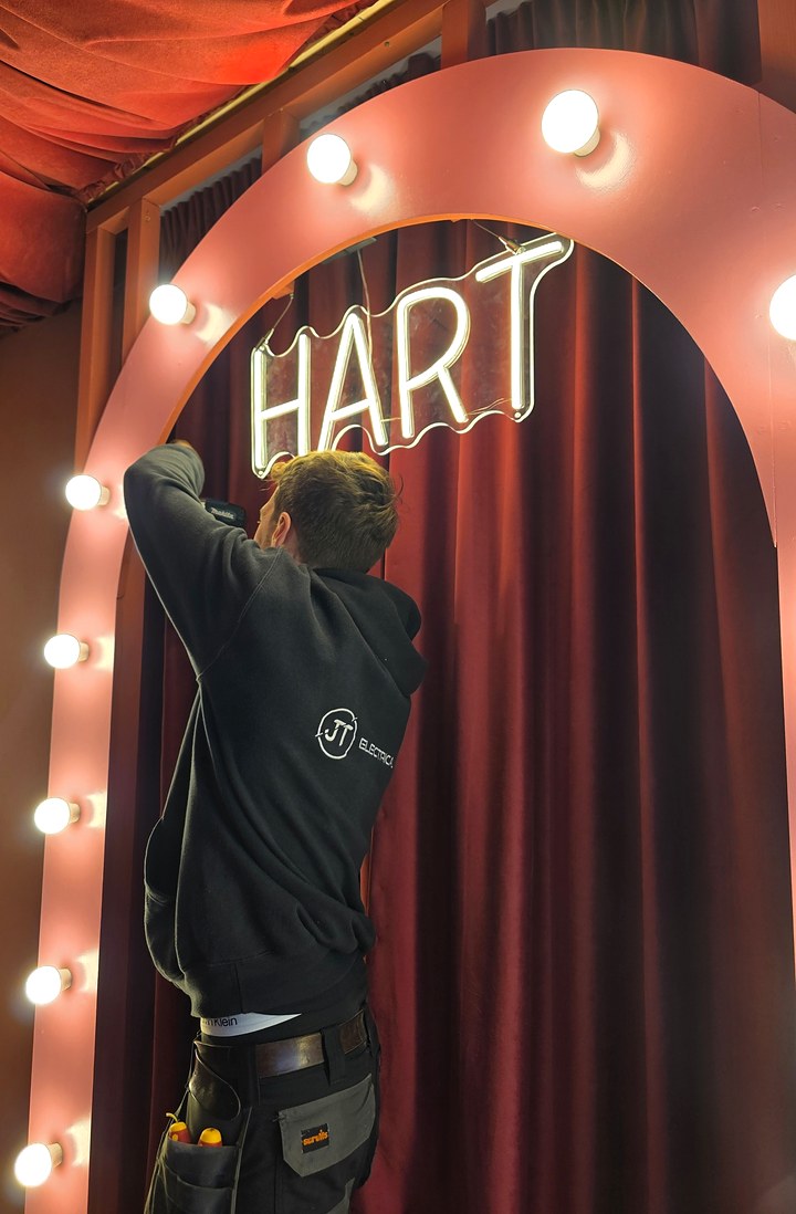 JT Electrical electrician in a branded hoodie fitting a neon sign inside an arch of festoon bulbs, against red velvet curtains