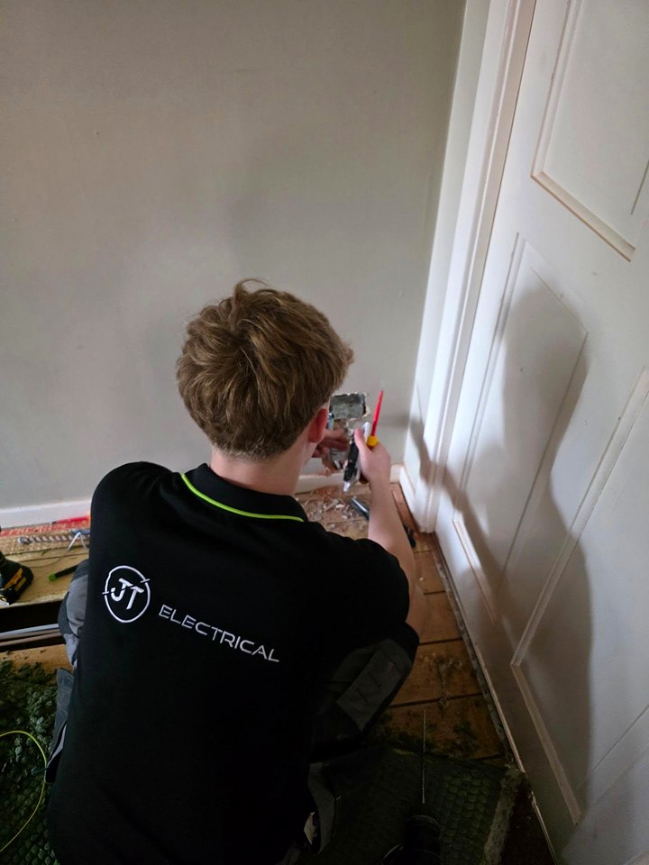 JT Electrical electrician in a branded black polo, seen from behind, wiring a socket at skirting level